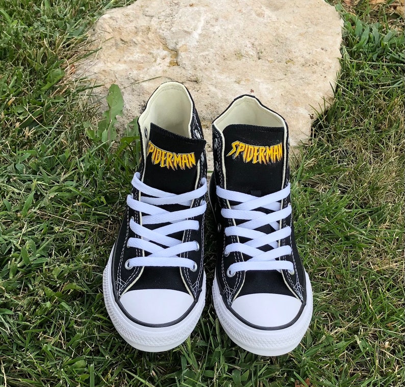 K&ouml;nnte beinhalten: Paar schwarze High-Top-Sneaker mit wei&szlig;en Schn&uuml;rsenkeln und Zehenkappen. Das Wort "SPIDERMAN" ist gelb auf der Zunge jedes Schuhs aufgedruckt. Die Schuhe stehen auf gr&uuml;nem Gras.