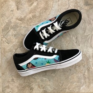 May include: Pair of black and blue Vans sneakers. The sneakers feature a light blue pattern with cartoon characters, white laces, and a white sole. The Vans logo is visible on the inner sole.
