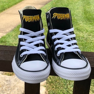 K&ouml;nnte beinhalten: Paar schwarze High-Top-Sneaker mit wei&szlig;en Schn&uuml;rsenkeln und einer wei&szlig;en Gummi-Zehenkappe. Das Wort "SPIDERMAN" ist goldfarben auf der schwarzen Zunge jedes Schuhs aufgedruckt. Die Schuhe werden auf einer Holzoberfl&auml;che mit gr&uuml;nem Gras im Hintergrund pr&auml;sentiert.
