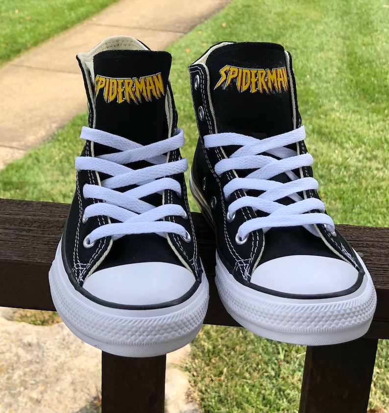 K&ouml;nnte beinhalten: Paar schwarze High-Top-Sneaker mit wei&szlig;en Schn&uuml;rsenkeln und einer wei&szlig;en Gummi-Zehenkappe. Das Wort "SPIDERMAN" ist goldfarben auf der schwarzen Zunge jedes Schuhs aufgedruckt. Die Schuhe werden auf einer Holzoberfl&auml;che mit gr&uuml;nem Gras im Hintergrund pr&auml;sentiert.