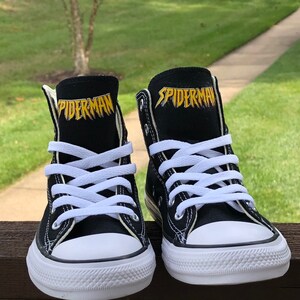 K&ouml;nnte beinhalten: Paar schwarze High-Top-Sneaker mit wei&szlig;en Schn&uuml;rsenkeln und Gummisohlen. Das Wort "SPIDERMAN" ist goldfarben auf der Zunge jedes Schuhs aufgedruckt. Die Schuhe stehen vor einem verschwommenen gr&uuml;nen Hintergrund.