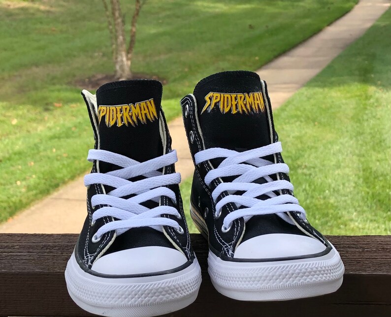 K&ouml;nnte beinhalten: Paar schwarze High-Top-Sneaker mit wei&szlig;en Schn&uuml;rsenkeln und Gummisohlen. Das Wort "SPIDERMAN" ist goldfarben auf der Zunge jedes Schuhs aufgedruckt. Die Schuhe stehen vor einem verschwommenen gr&uuml;nen Hintergrund.