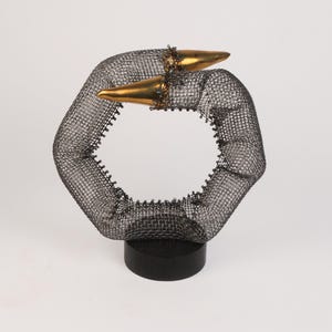 Puede incluir: Una escultura de alambre con forma circular y dos puntas doradas. La escultura está sobre una base de madera negra.