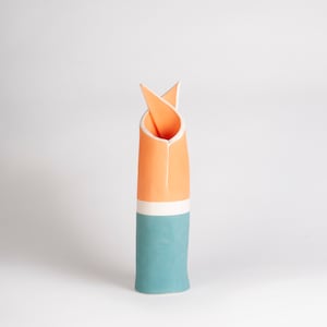 Puede incluir: Un jarrón de cerámica alto con un diseño plegado único. El jarrón presenta un diseño de bloques de color con una base turquesa, una franja blanca y una sección superior naranja. El interior es naranja. La parte superior está doblada.