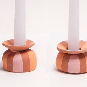 May include: Two ceramic candle holders with a striped design in shades of orange and pink. Each holder has a circular base and a top to hold a white candle. The candle holders are a decorative home accent.