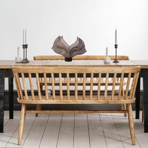 May include: A dining room scene featuring a wooden table with black legs, a matching bench, and two chairs. The table is set with candles in black metal holders and a decorative sculpture. The room has a neutral color palette.