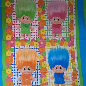 May include: A colorful fabric panel featuring four Troll dolls with vibrant hair in green, pink, orange, and blue. Each doll is surrounded by a floral border with a white polka dot background. A measuring tape is on the right side.