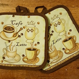 May include: Two brown and beige pot holders with a coffee theme. The pot holders feature illustrations of coffee cups and the words "Cafe" and "Latte".