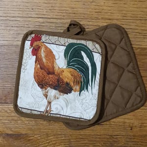 May include: Two brown quilted pot holders. One pot holder features a colorful rooster illustration with a red comb, brown and yellow feathers, and a green tail. The other pot holder is solid brown.