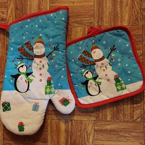 May include: A set of two blue and red Christmas oven mitts with a snowman and penguin design. The mitts are decorated with snowflakes and stars.