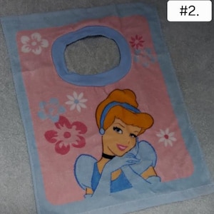 May include: Pink and blue baby bib featuring a Cinderella graphic. The bib has a blue border and a circular opening at the top. Floral designs are scattered around the Cinderella image. The text "#2." is in the top right corner.