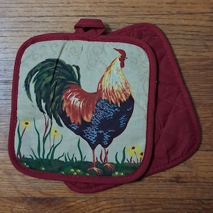 May include: A square quilted potholder featuring a central illustration of a rooster with red, orange, and dark blue feathers. The background is a light cream color with faint scrollwork, green grass, and small yellow flowers. It has a red fabric border.