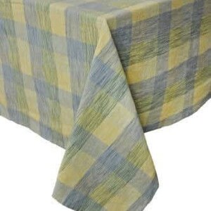 May include: A close-up of a tablecloth with a plaid pattern in shades of yellow, blue, and green. The fabric is draped over a surface, with a corner of the cloth hanging down. The tablecloth has a textured appearance.