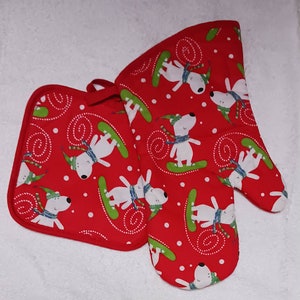 May include: Red oven mitts with a white dog wearing a scarf and riding a snowboard pattern. The mitts are made of cotton and have a loop for hanging.