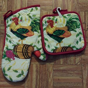 May include: Oven mitt and pot holder set with a rooster design. The white fabric features a colorful rooster standing on a wooden barrel, surrounded by green leaves and the words "Good morning" and "Apple". The set is trimmed with red fabric.