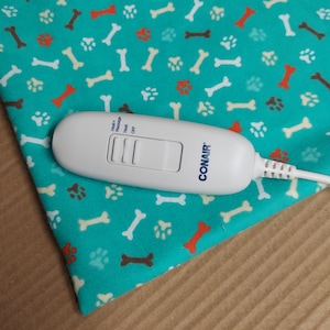 May include: White electric heating pad controller with a blue and white paw print design. The controller has buttons for heat, massage, and off. The text "CONAIR" is printed on the controller.