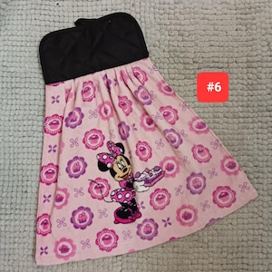 May include: A hanging kitchen towel with a black quilted top and a light pink fabric bottom featuring a repeating pattern of purple cupcakes and a graphic of Minnie Mouse holding a tray of cupcakes. The number 6 is printed in a red square.