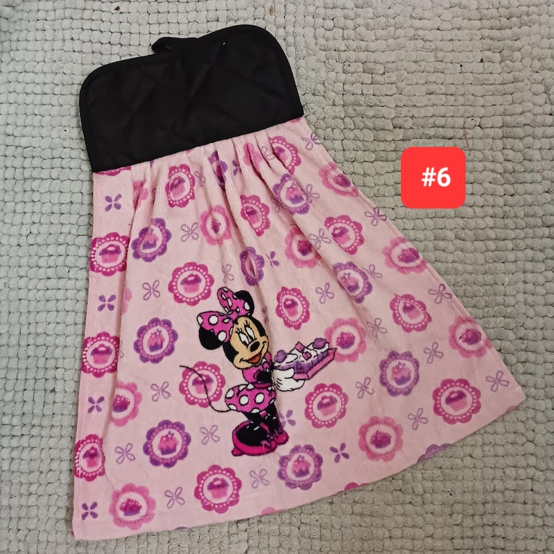 May include: A hanging kitchen towel with a black quilted top and a light pink fabric bottom featuring a repeating pattern of purple cupcakes and a graphic of Minnie Mouse holding a tray of cupcakes. The number 6 is printed in a red square.
