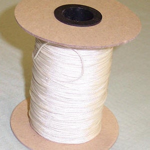 May include: A spool of off-white thread. The thread is tightly wound around a cardboard core with a black center. The spool has two large, round cardboard flanges on the top and bottom.