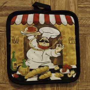 May include: A black and white pot holder with a cartoon chef holding a plate of spaghetti and a meatball on a skewer. The chef is wearing a white chef's hat and a red bandana. The background is a yellow and brown building with a red and white awning. The pot holder has a black border.