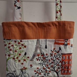 May include: A fabric wall hanging pocket with two top straps, featuring a white background with a printed scene of the Eiffel Tower, a bicycle, trees with red flowers, and an orange building. The top section is a solid orange fabric.