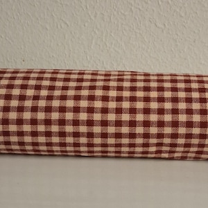 May include: A long, cylindrical pillow with a red and cream checkered pattern. The fabric is tightly woven, and the pillow appears to be stuffed. The pillow is likely used for decorative or functional purposes.