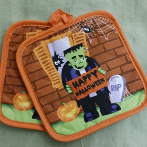 May include: A pair of orange and green Halloween themed oven mitts. The mitts feature a brick wall with a window, a Frankenstein monster holding a sign that says "Happy Halloween", a spider web, a tombstone that says "RIP", and pumpkins.