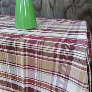 May include: A red, brown, and beige plaid tablecloth with a white background. The tablecloth is made of a woven fabric and has a slightly textured surface.