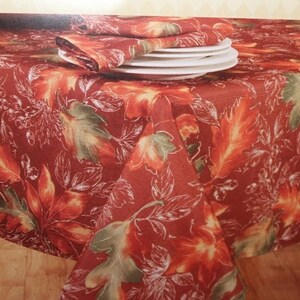 May include: Red tablecloth with a white and green leaf pattern. The tablecloth is laid on a wooden table with white plates and napkins on top.