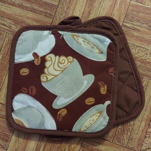 May include: A pair of brown quilted pot holders with a brown trim. The front of the pot holder features a repeating pattern of coffee cups with whipped cream and coffee beans on a brown background.
