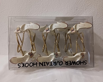 May include: Set of six white shower curtain hooks with gold rings and butterfly accents. The hooks are packaged in a clear plastic box with the words "SHOWER CURTAIN HOOKS" printed on the bottom.