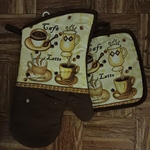 May include: A set of two brown and beige kitchen pot holders with a coffee and latte theme. The pot holders feature illustrations of coffee cups and the words "Cafe" and "Latte".