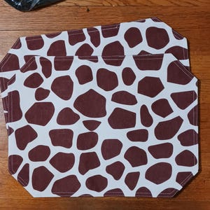 May include: Two rectangular placemats with a giraffe print pattern. The placemats feature a white background with brown irregular shapes, resembling a giraffe's coat. The edges are neatly stitched.