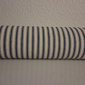 May include: A cylindrical bolster pillow with a cream-colored background and vertical navy blue stripes. The pillow is made of fabric and appears to be a decorative item for a bed or sofa.