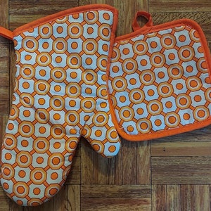 May include: A set of two orange and white patterned oven mitts. The larger mitt is a potholder with a hanging loop. The smaller mitt is a standard oven mitt.