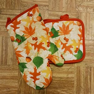 May include: An oven mitt and pot holder set with a fall-themed leaf pattern. The mitt and pot holder are cream-colored with orange, yellow, and green leaves, acorns, and berries. The edges are trimmed with red fabric.