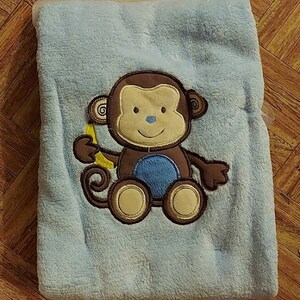 May include: A light blue baby blanket with an embroidered monkey holding a banana. The monkey is brown with blue eyes and a yellow banana.