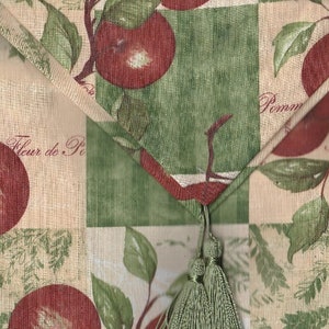 May include: Close-up of a decorative fabric with a repeating apple and leaf pattern. The design features red apples, green leaves, and the words "Fleur de Pom" and "Pommes" in script. The fabric has a beige and green color scheme.