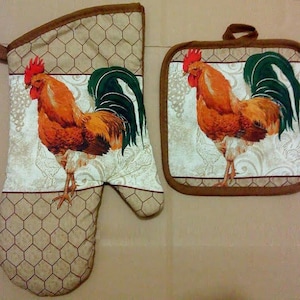 May include: A set of two brown and beige oven mitts with a rooster design. The rooster is brown, red, and green with a white background. The mitts have a brown border and a chicken wire pattern.