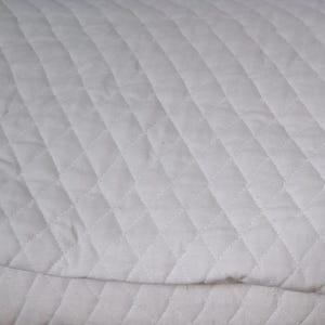 May include: Close-up of a white quilted fabric with a diamond pattern. The fabric appears soft and has a subtle texture, with visible stitching outlining each diamond shape. The overall look is clean and simple.