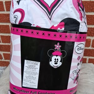 May include: A Disney Minnie Mouse microfibre full comforter packaged in plastic. The comforter features a white, pink, and black heart pattern. The packaging label displays the Disney logo, a Minnie Mouse graphic, and text stating 193cm x 218cm.