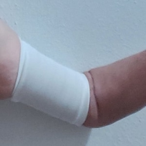 May include: White elbow support made of soft fabric. The support is worn on the left arm.