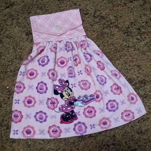 May include: A pink hanging kitchen towel featuring a repeating pattern of purple cupcakes in scalloped frames and a central graphic of Minnie Mouse holding a tray of treats. The top section has a light pink fabric with a white geometric pattern.