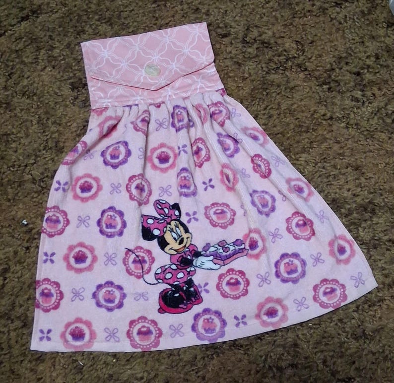 May include: A pink hanging kitchen towel featuring a repeating pattern of purple cupcakes in scalloped frames and a central graphic of Minnie Mouse holding a tray of treats. The top section has a light pink fabric with a white geometric pattern.