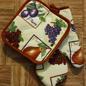 May include: A pair of oven mitts with a white background and brown trim. The mitts feature a colorful fruit and floral design with plums, pears, and grapes. The text "Plums" and "Pear" is printed on the mitts.