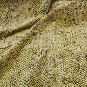 May include: Close-up of a fabric with a snakeskin pattern. The fabric has a tan background with dark brown spots and a textured appearance. The material is draped, showing folds and shadows.