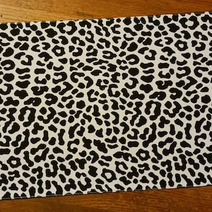 May include: Black and white leopard print placemat. The placemat is made of a fabric material and has a black border.