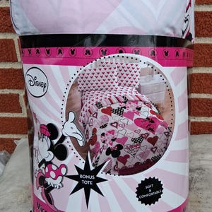 May include: A Disney Minnie Mouse microfibre full comforter set in packaging. The bedding features a pink, white, and black heart and character pattern. The packaging indicates a 193 cm by 218 cm size, includes a bonus tote, and is labelled as soft and comfortable.