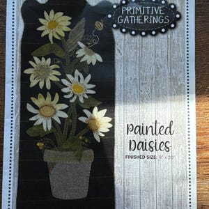 May include: A sewing pattern sheet titled Primitive Gatherings Painted Daisies with a finished size of 9 inches by 20 inches. The design features white daisies with yellow centers in a grey pot on a black background with a small bee illustration.