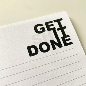May include: White notepad with the words "GET IT DONE" printed in bold black letters. The notepad has horizontal lines for writing and is set against a light background.